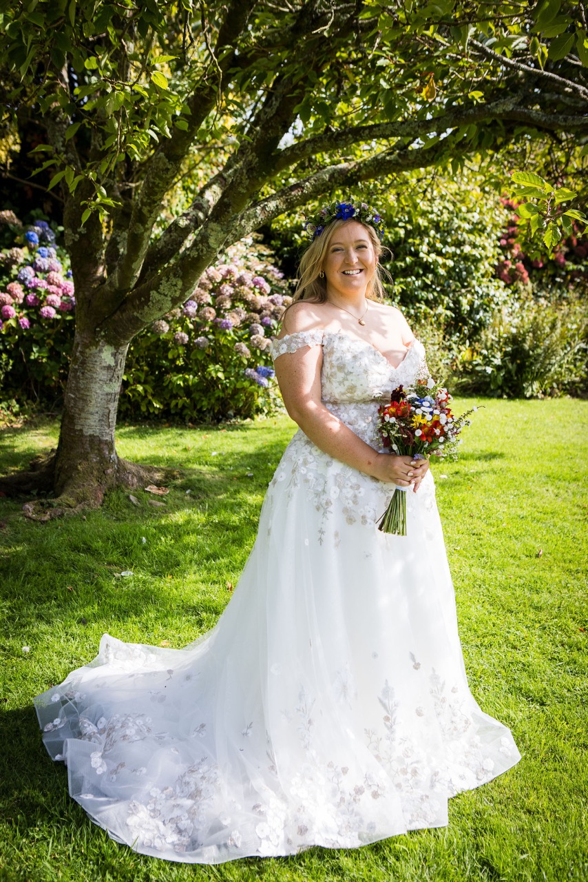bride wearing a flower crown and holding a colourful bouquet, standing beneath a tree