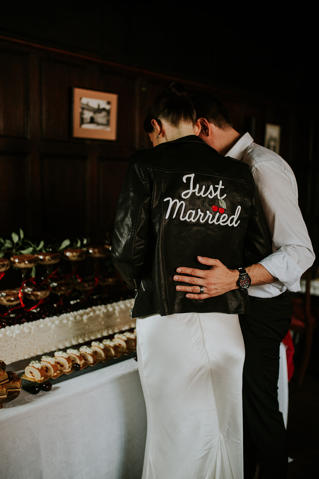 bride and groom face away from the camera looking at the wedding breakfast desert table. The bride wears a 'just married' leather jacket with cherries on it