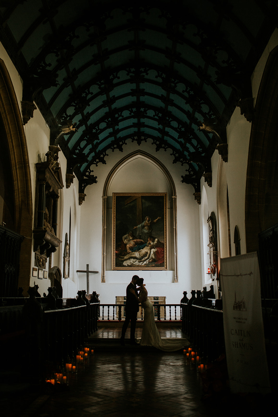 artistic photo of hte inside of a church where the front of the church is lit and the bride and groom are silhouetted. Candles light the aisle