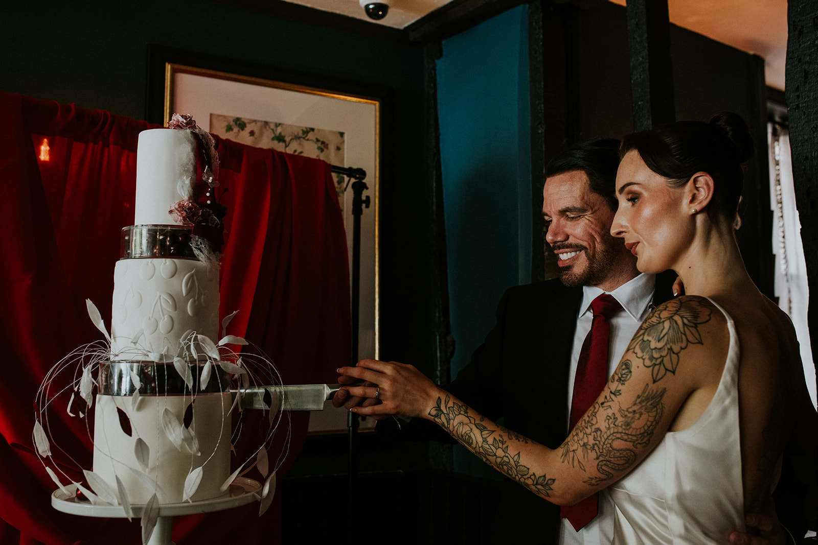 bride and groom cut the base layer of their sculptural wedding cake with embossed cherries as part of their cherry red winter wedding
