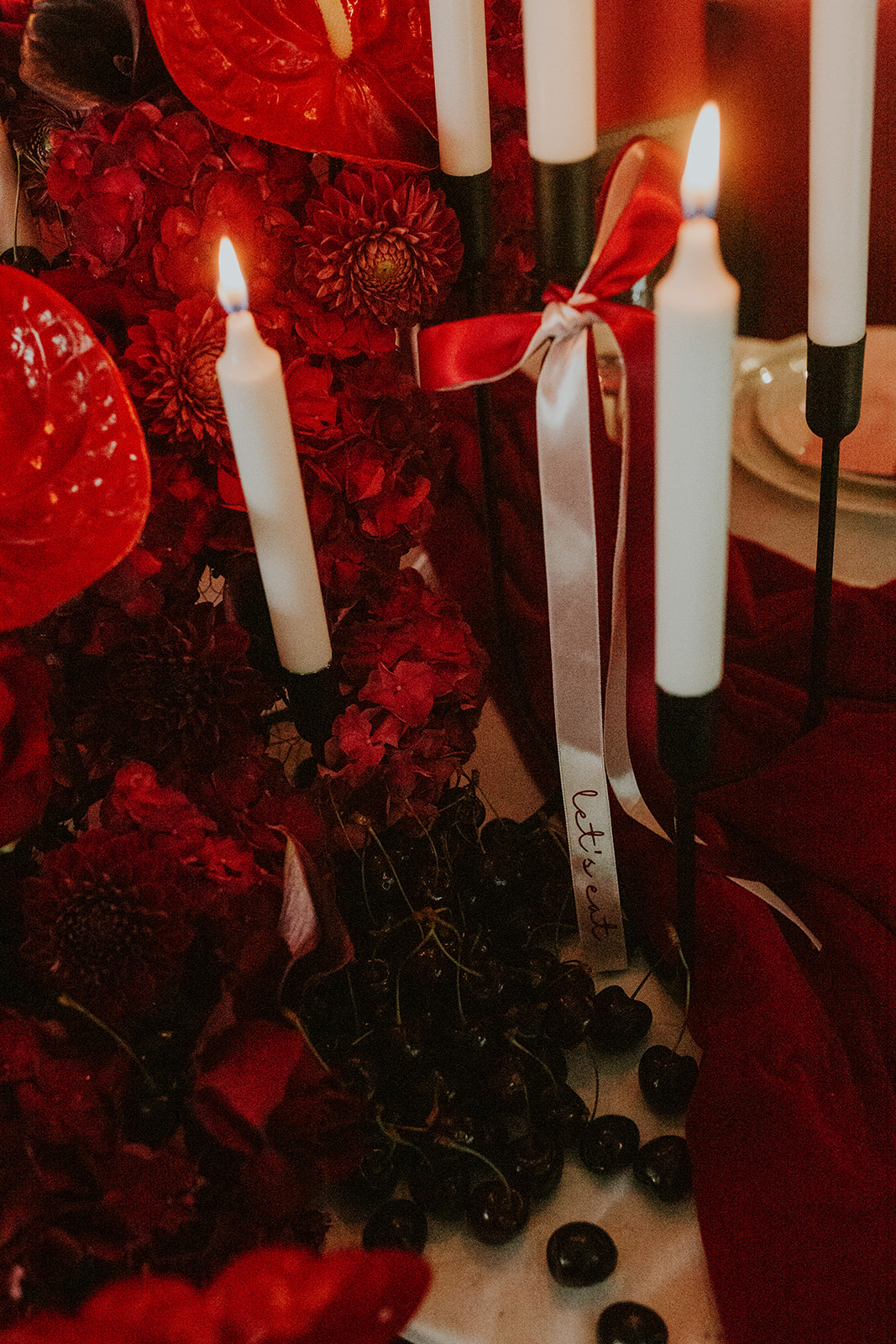detail shot of a ribbon with 'lets eat' written on it. It is tied around a candelabra which is surrounded with red flowers dor cherry red winter wedding
