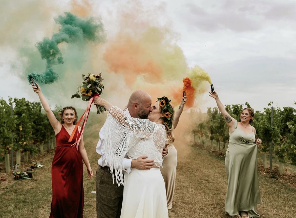 bride and groom kissing in a field of grape vines while bridesmaids hold coloured smoke bombs