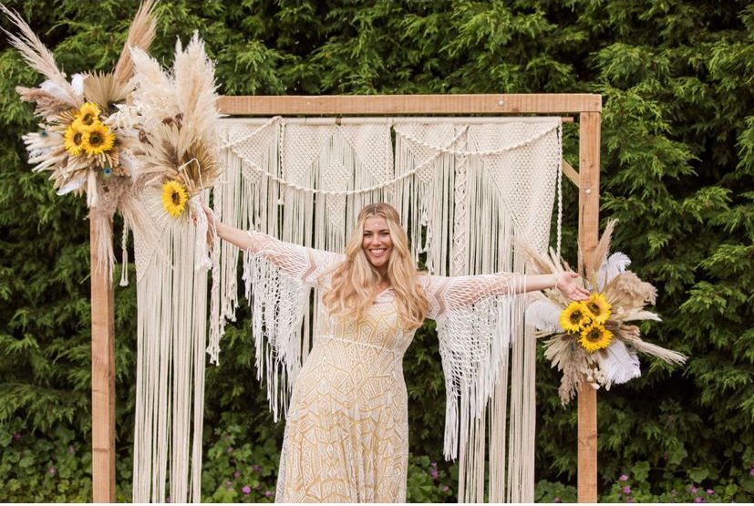 bride wearing tassel sleeved dress and head chain stood in front of macrame backdrop as part of festival wedding