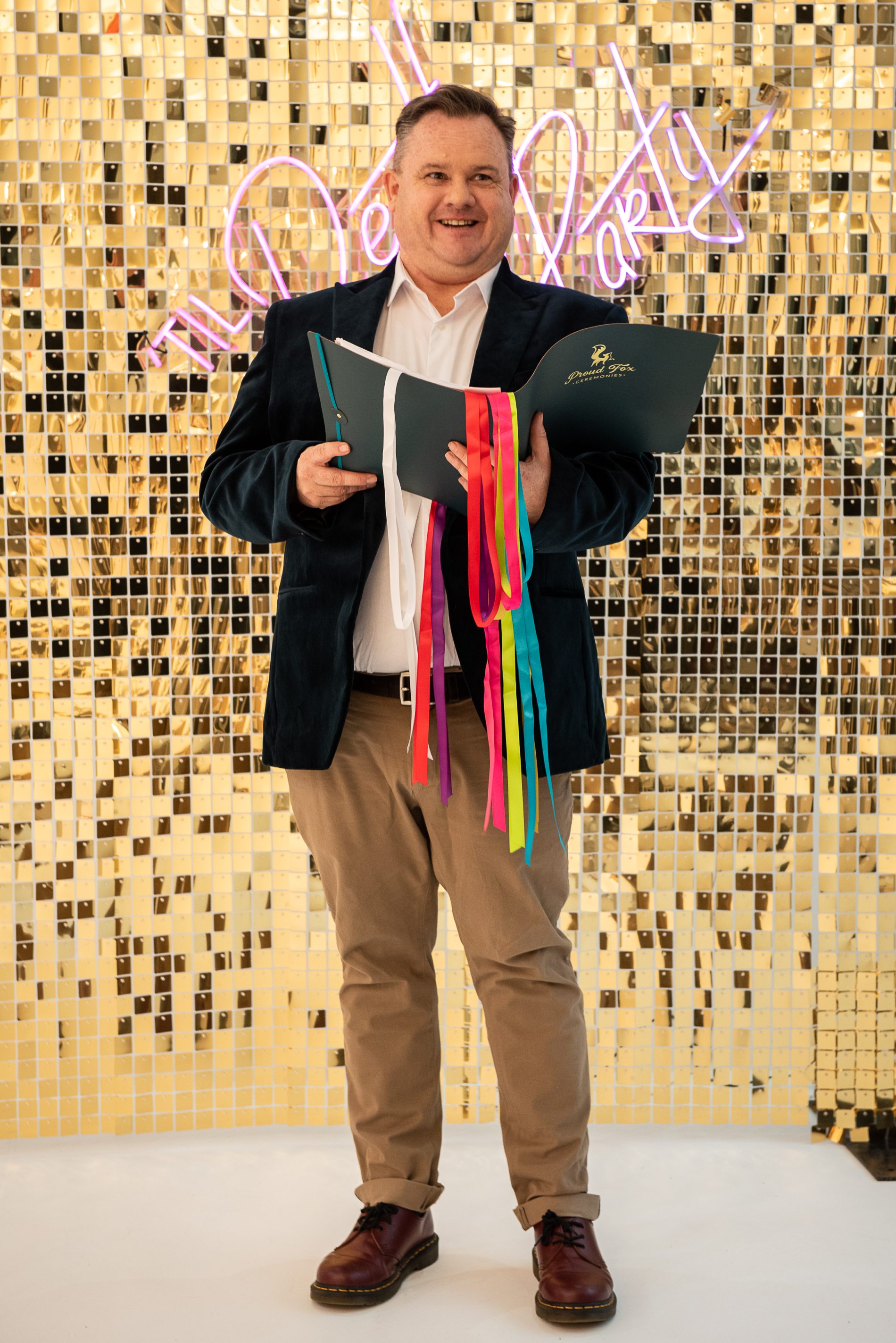 Proud Fox ceremonies stood holding ceremony book in front of glitter wall.