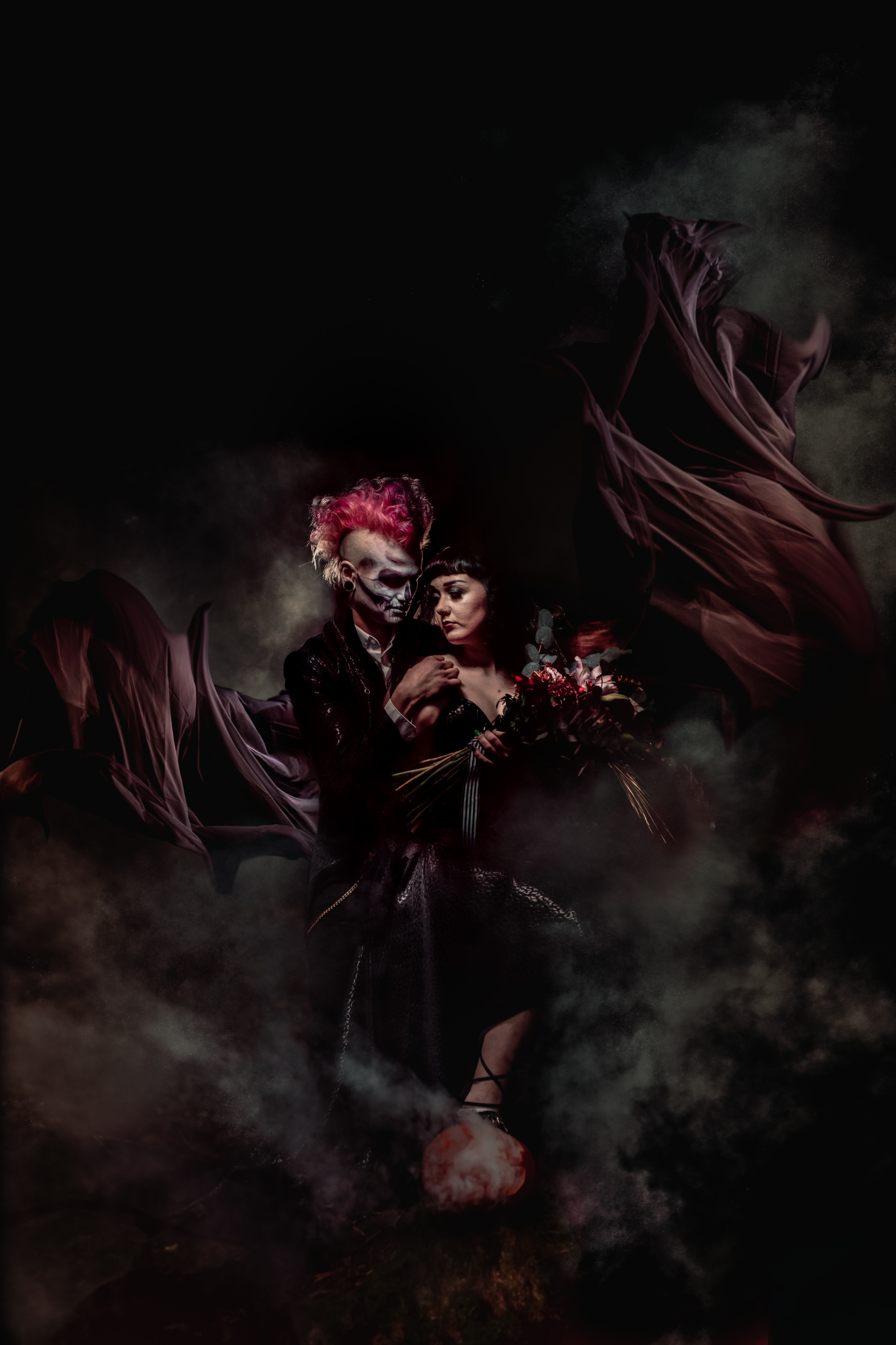dramatic photo of halloween wedding couple surrounded by black veils swirling in the wind - groom with dramatic pink mohawk and painted face - alternative bride hold bold wedding bouquet