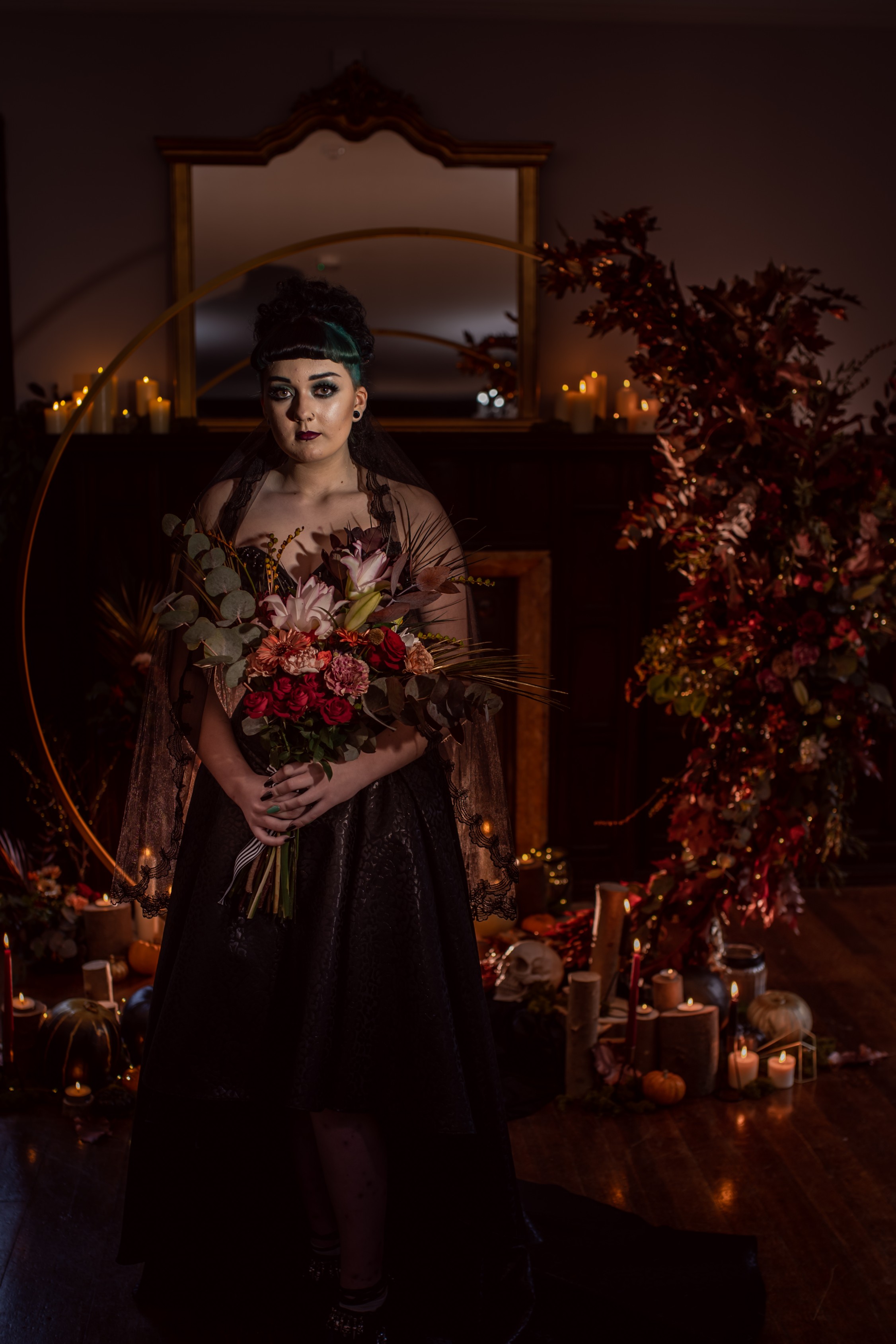 halloween bride - gothic halloween wedding set up - bride standing in front of dramatic floral arch in the candlelight with wedding bouquet - autumn colours of gold, copper and black