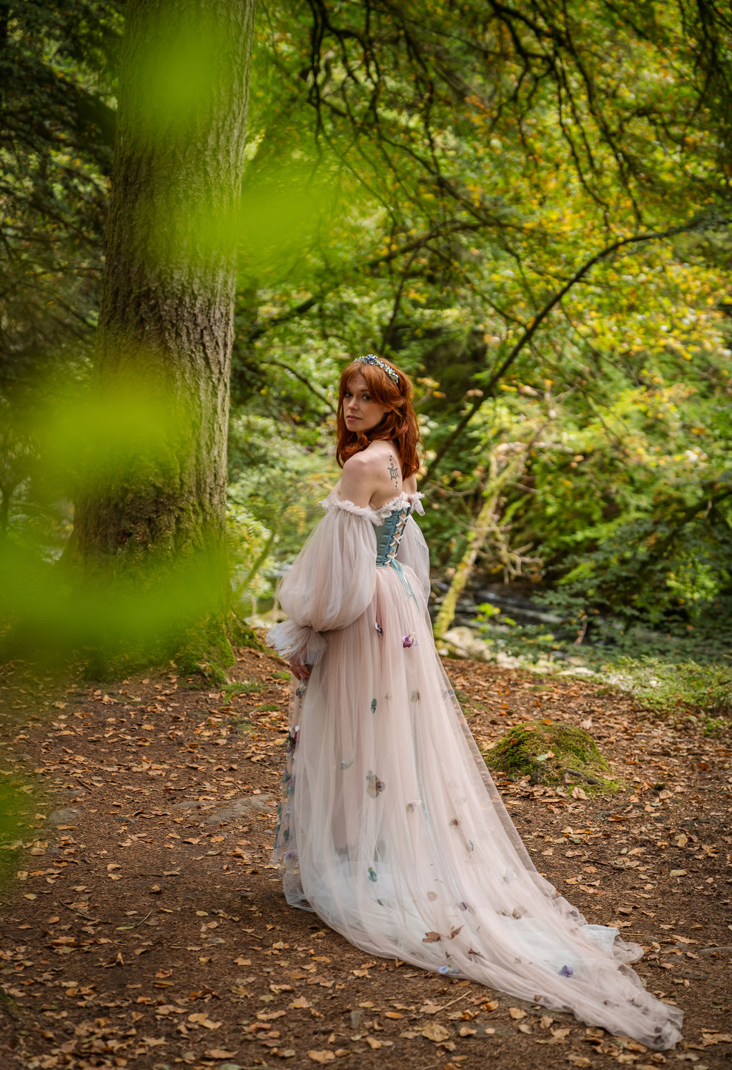 Bride stands in a forest wearing a off white whimsical wedding dress that has a blue corset style boddice. Bride has beautiful long auburn hair.