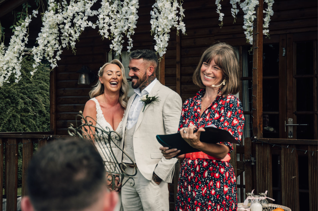 bride and groom stood under a white wisteria archway. groom wears a white suit.