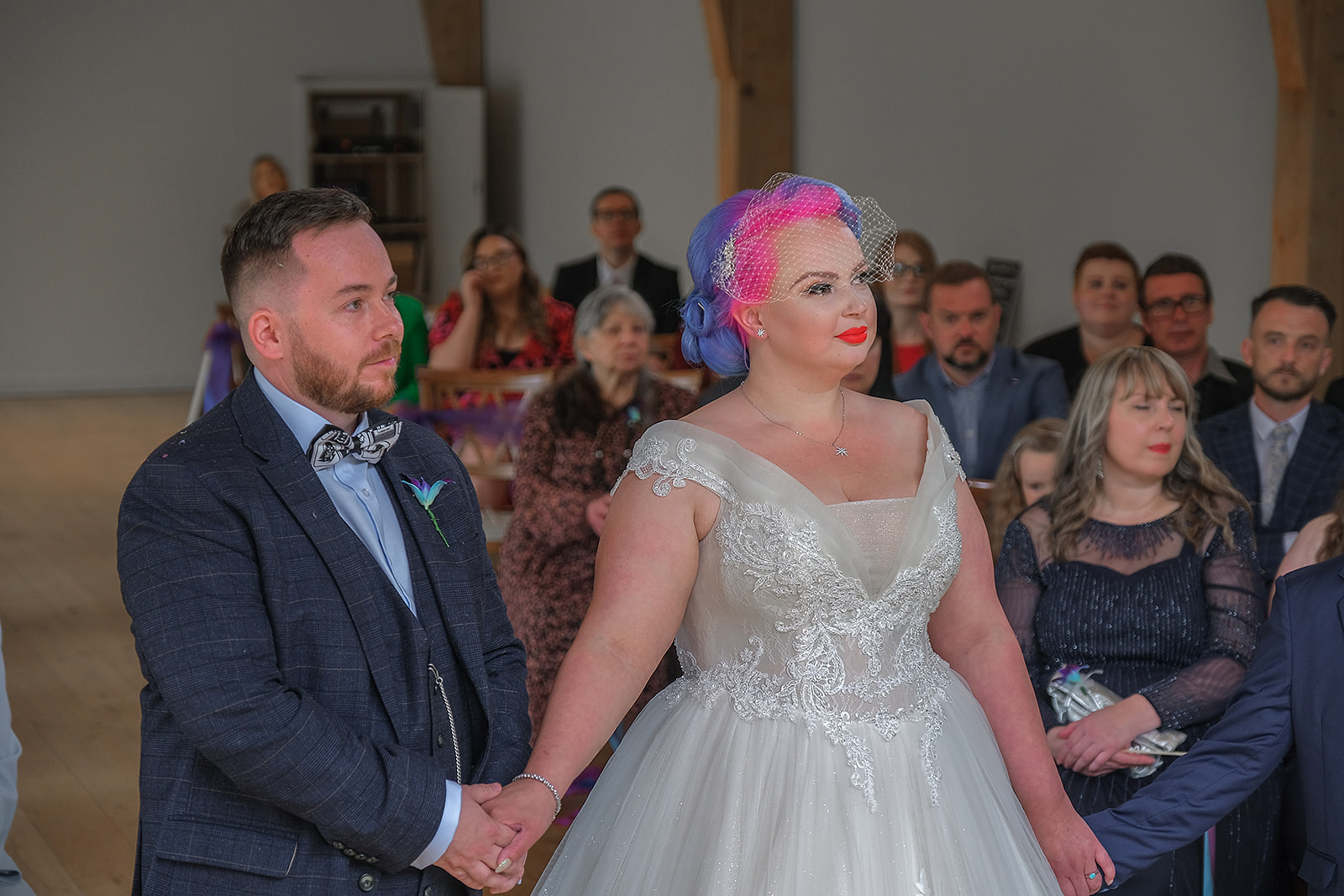 bride with pink hair and white voluminous dress, holding hands with groom in plaid suit and bow tie