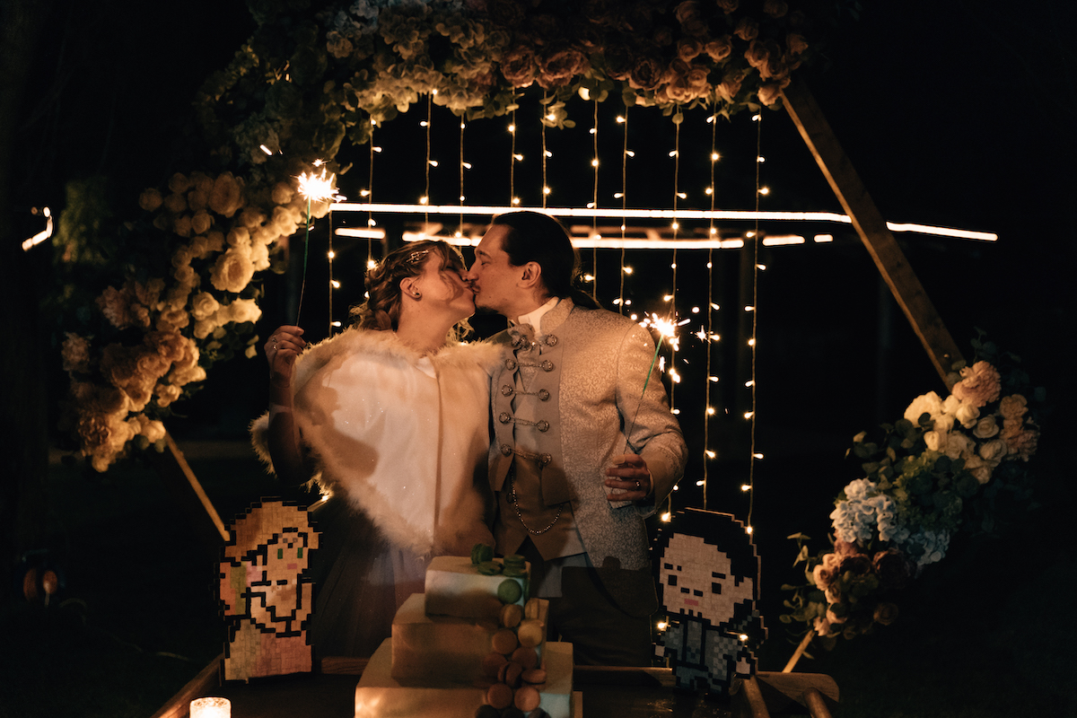 bride and groom kiss under hexagonal floral arch. handmade pixel bride and groom sit either side of pixel inspired wedding cake