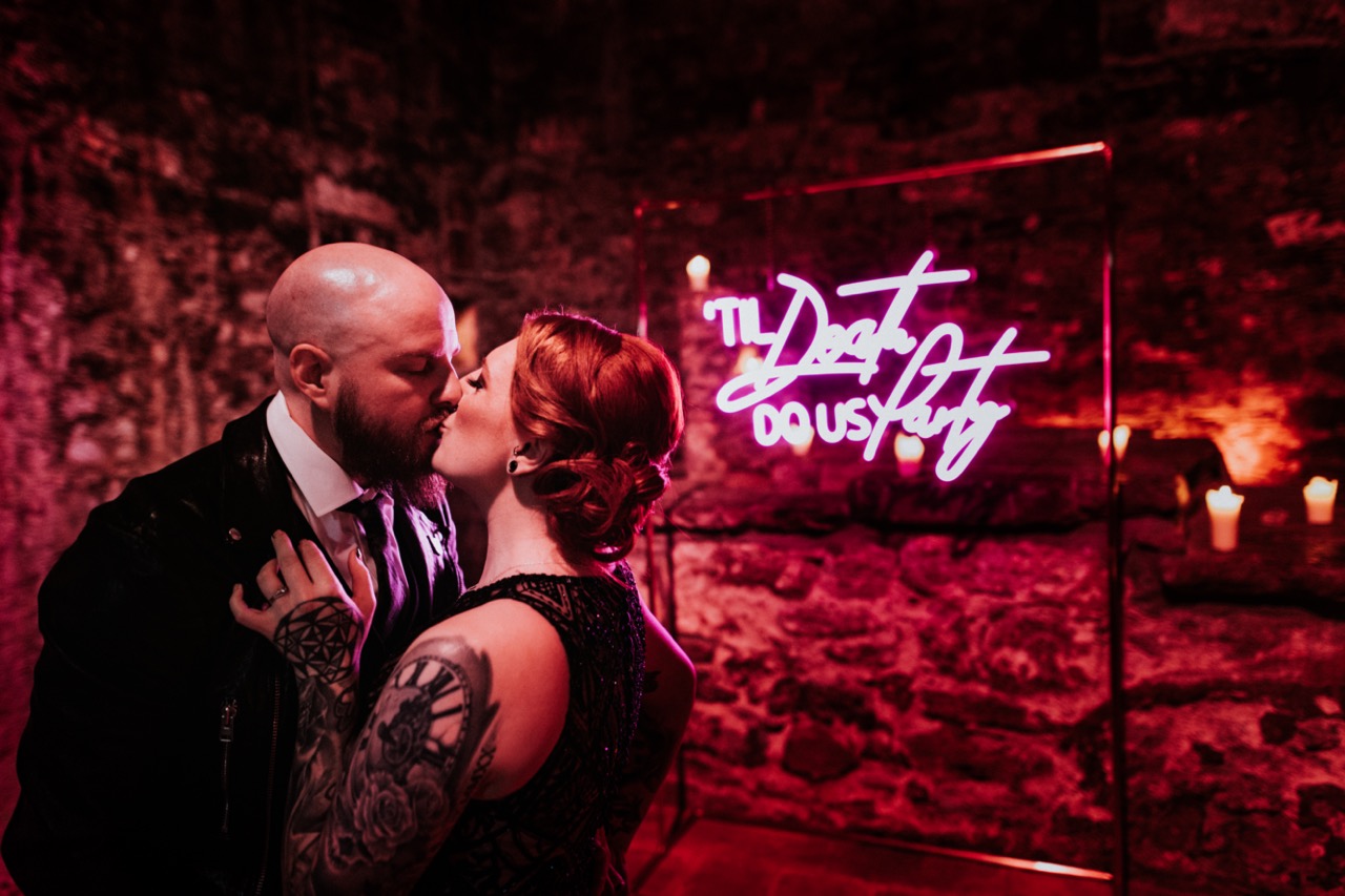 bride and groom kissing next to neon 'til death do us party' sign - unique wedding neon sign