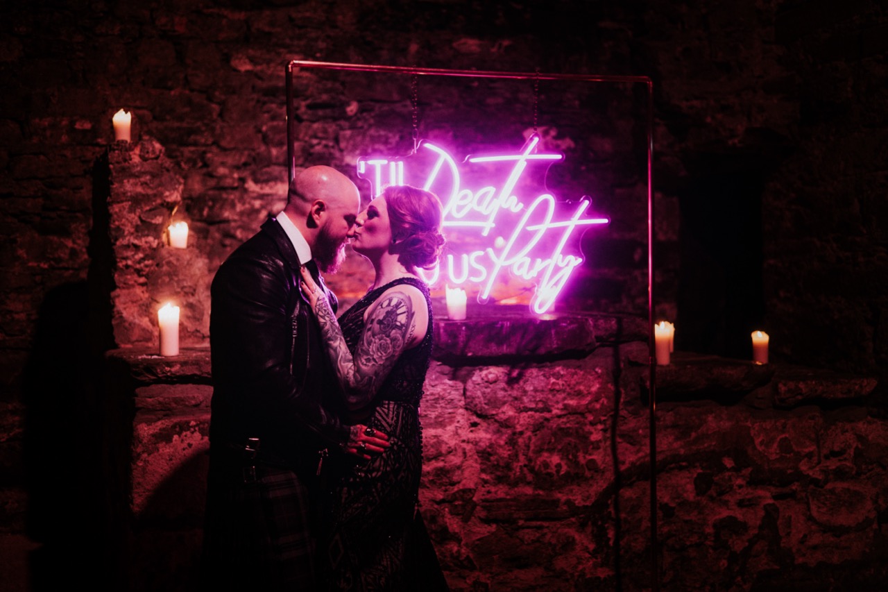 bride and groom kissing next to neon 'til death do us party' sign - unique wedding neon signage
