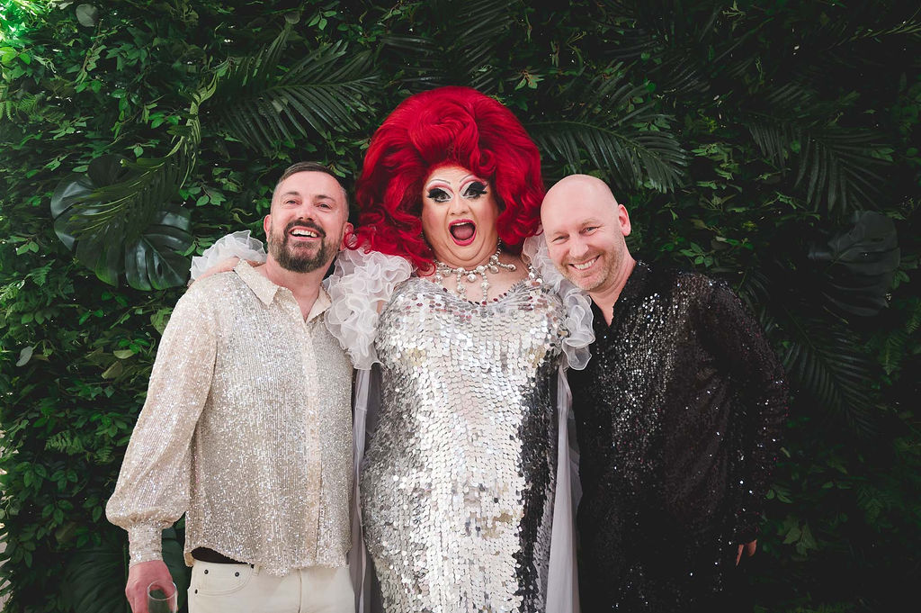 two grooms with their drag queen celebrant, all wearing sparkly shirts and smiling