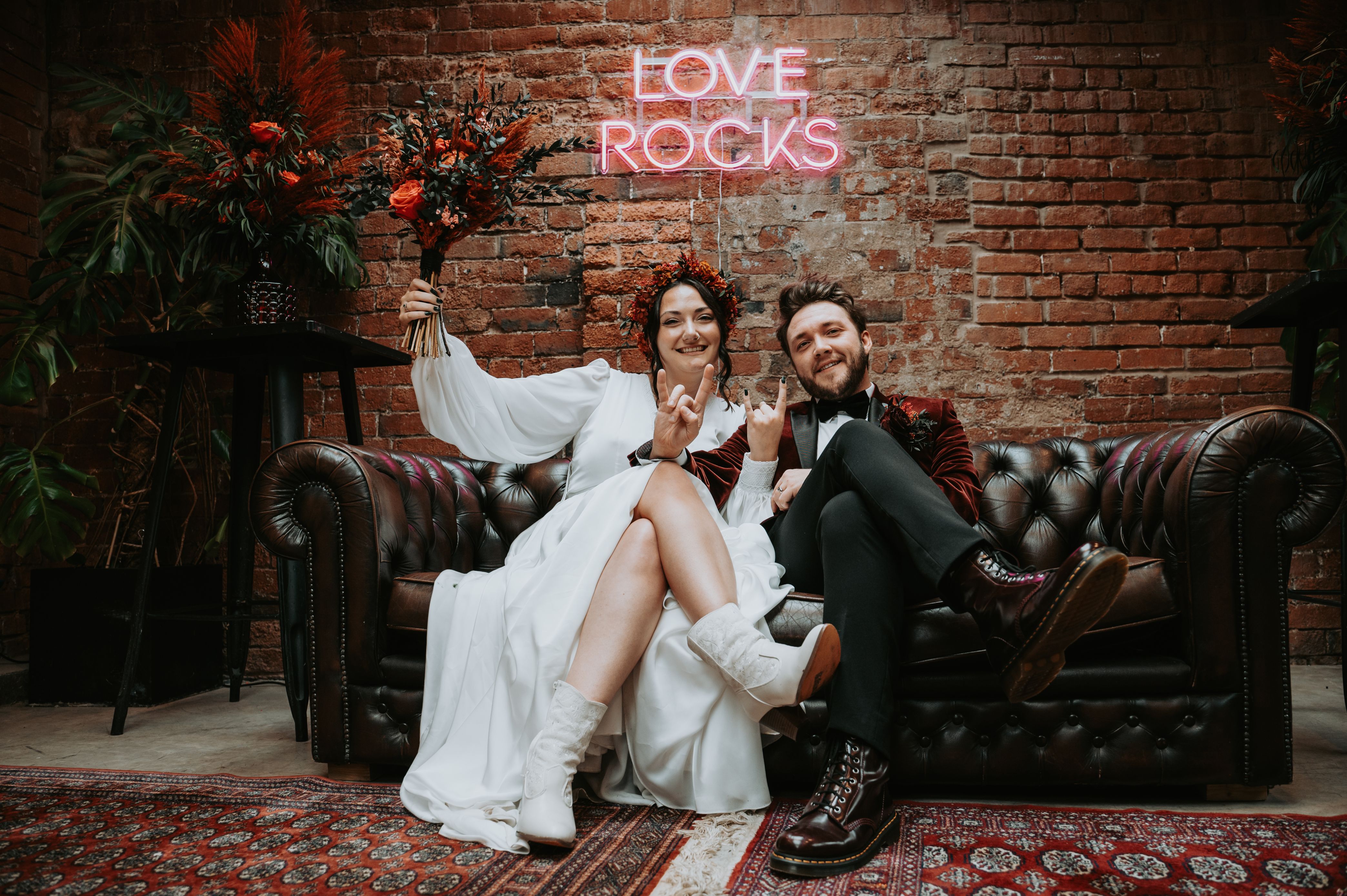 newlyweds sat together on a black leather sofa at the shack revolution industrial wedding venue in hereford with a neon love rocks sign behind