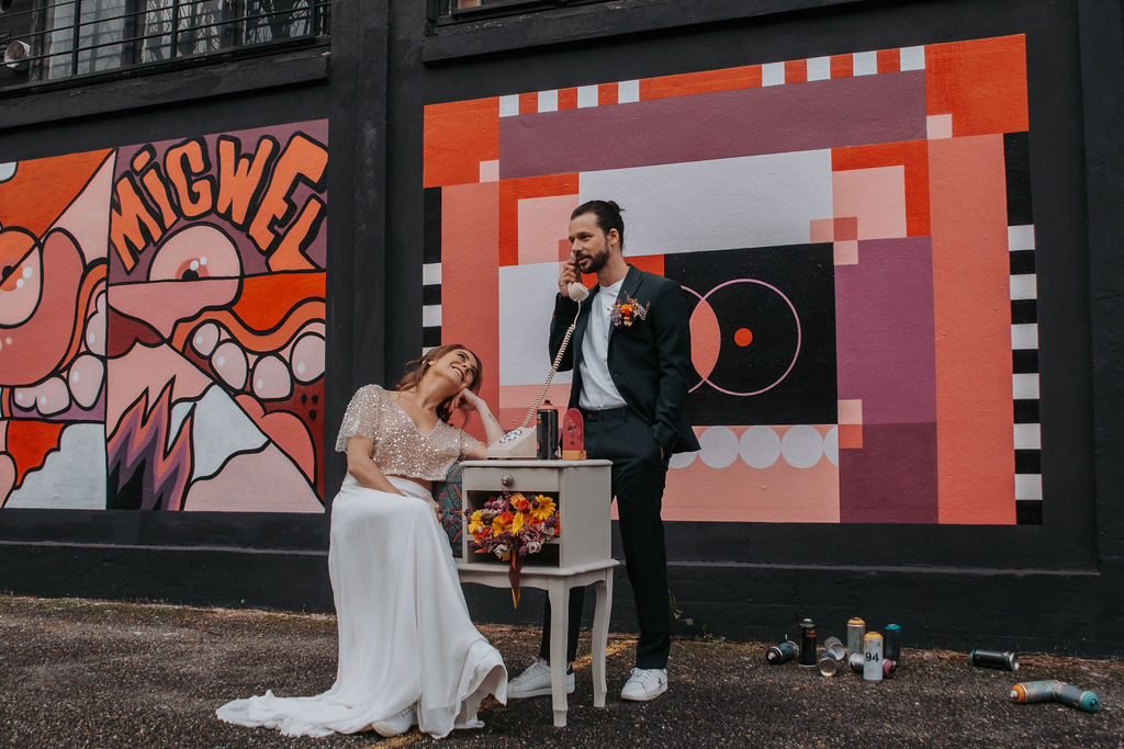 alternative bride and groom stand in front of a graffiti wall and play with a old fashioned telephone