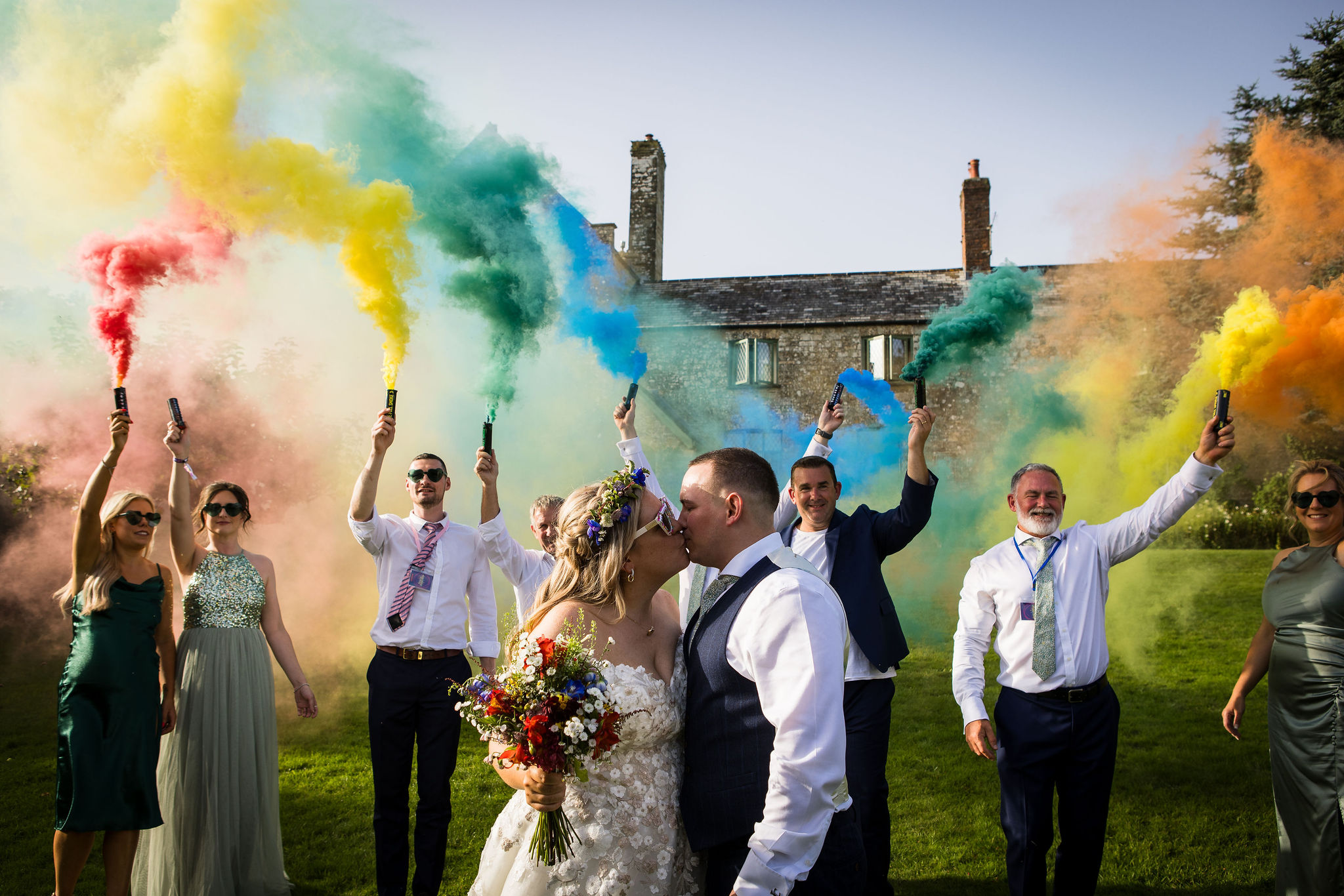 bride and groom kiss as their family hold rainbow smoke bombs