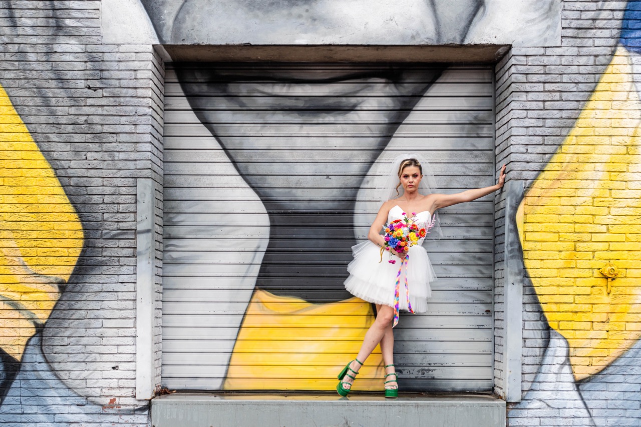 bride wears mini length wedding dress and green platform bridal shoes, has a multicoloured wedding bouquet and stands in front of a graffiti wall.