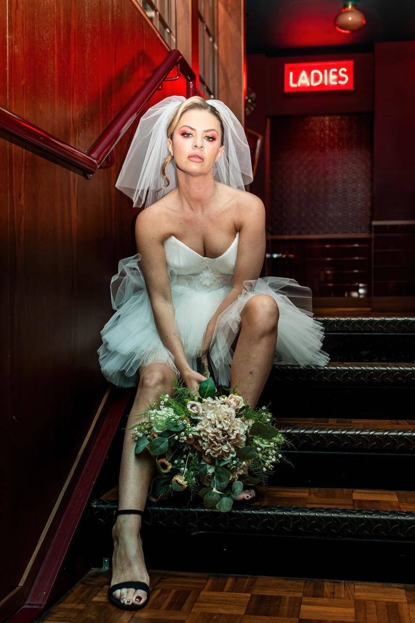 bride wears a mini length wedding dress and sits in a dark stairwell under a neon red light