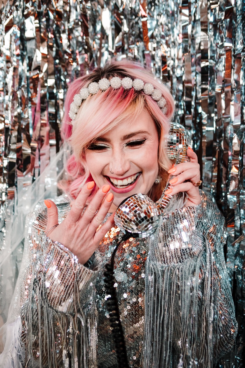 sparkly wedding ideas - bride wearing silver sparkly jacket speaking into disco ball audio guestbook