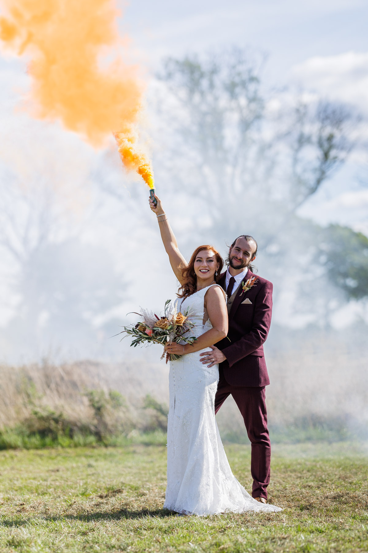 outdoor wedding photo bride and groom smiling and holding orange smoke bomb