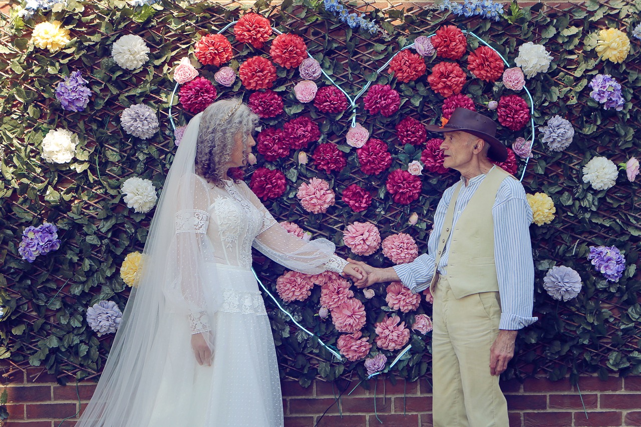 older bride and groom holding hands in front of flower wall