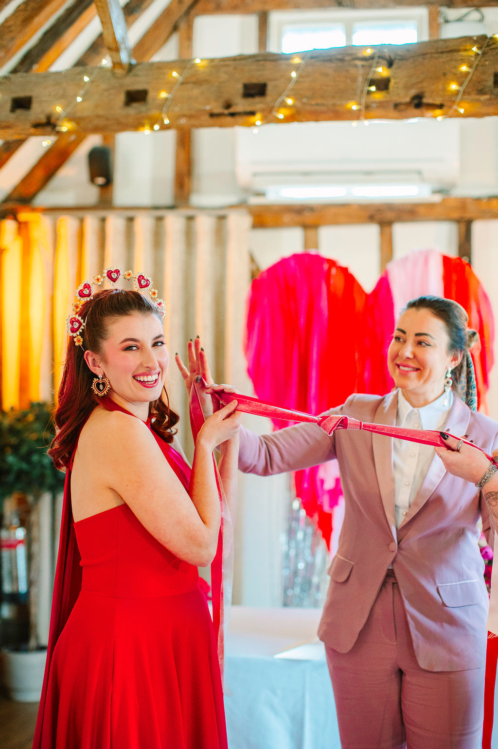 Bride and celebrant holding pink and red hand fasting ribbons. Pink love heart ceremony backdrop in the background.