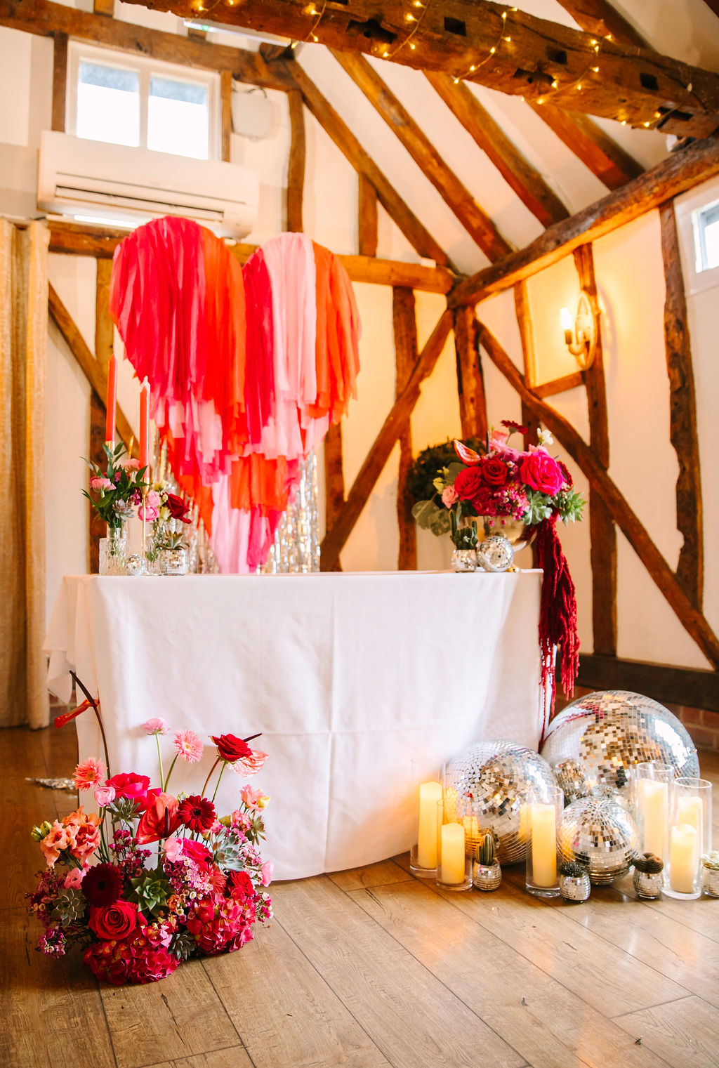 Beamed rustic wedding venue with fun Valentines wedding ceremony set up. Pink and red tassel heart with silver streamer focal point. Red and pink wedding flowers are set on the table and a display of wedding flowers on the floor. Disco balls and candles are to the right of the table.