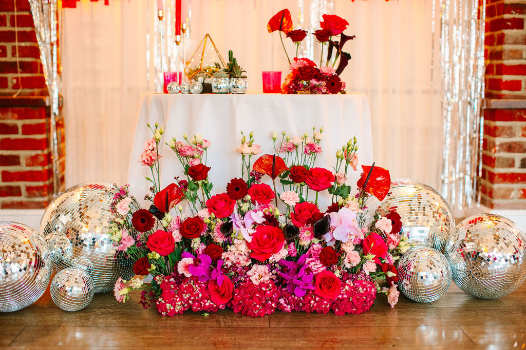Couple's sweetheart table with vibrant pink and red wedding flower display and disco balls for fun Valentines wedding