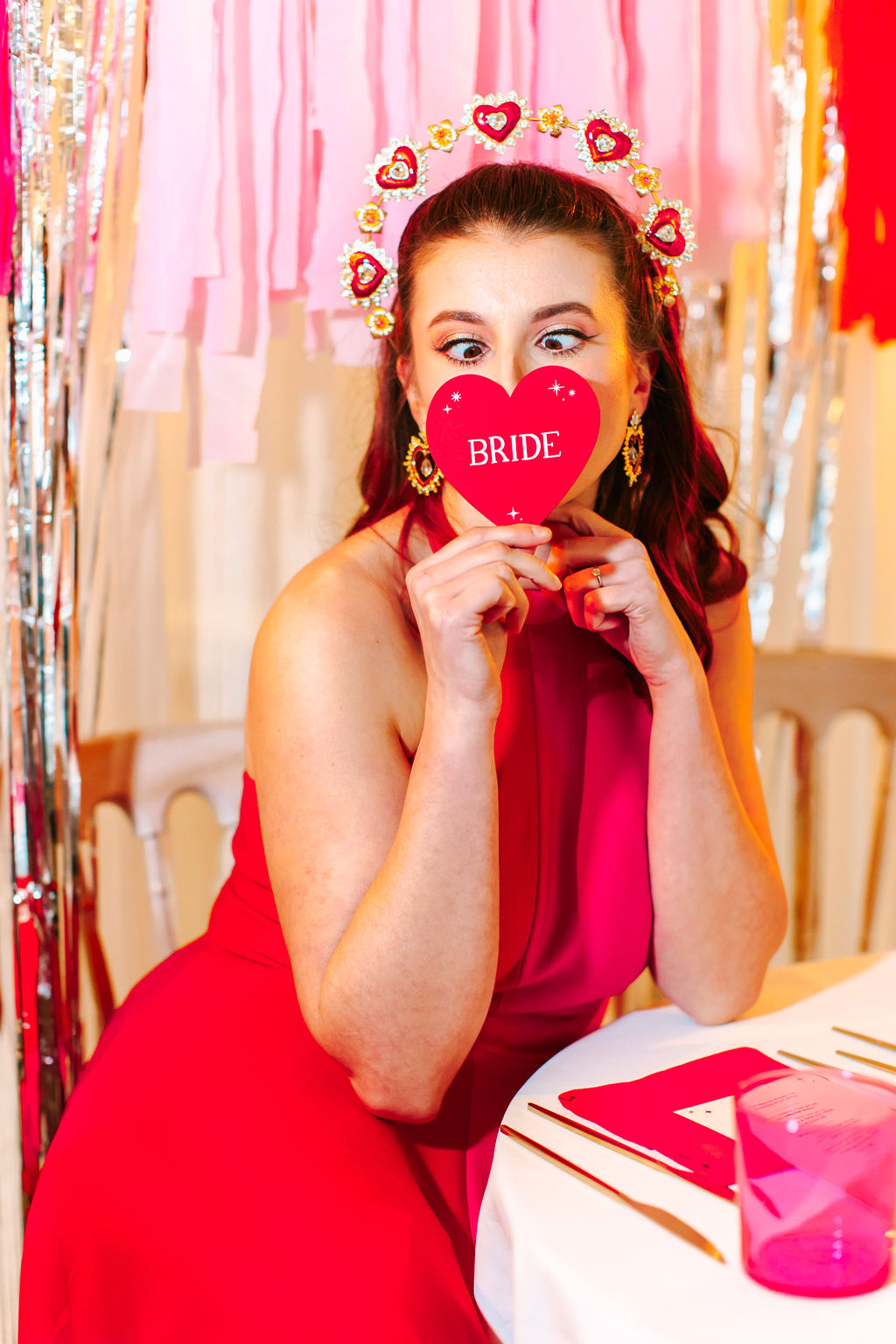 Bride holding 'bride' wedding place setting in front of her face at fun red and pink love heart wedding