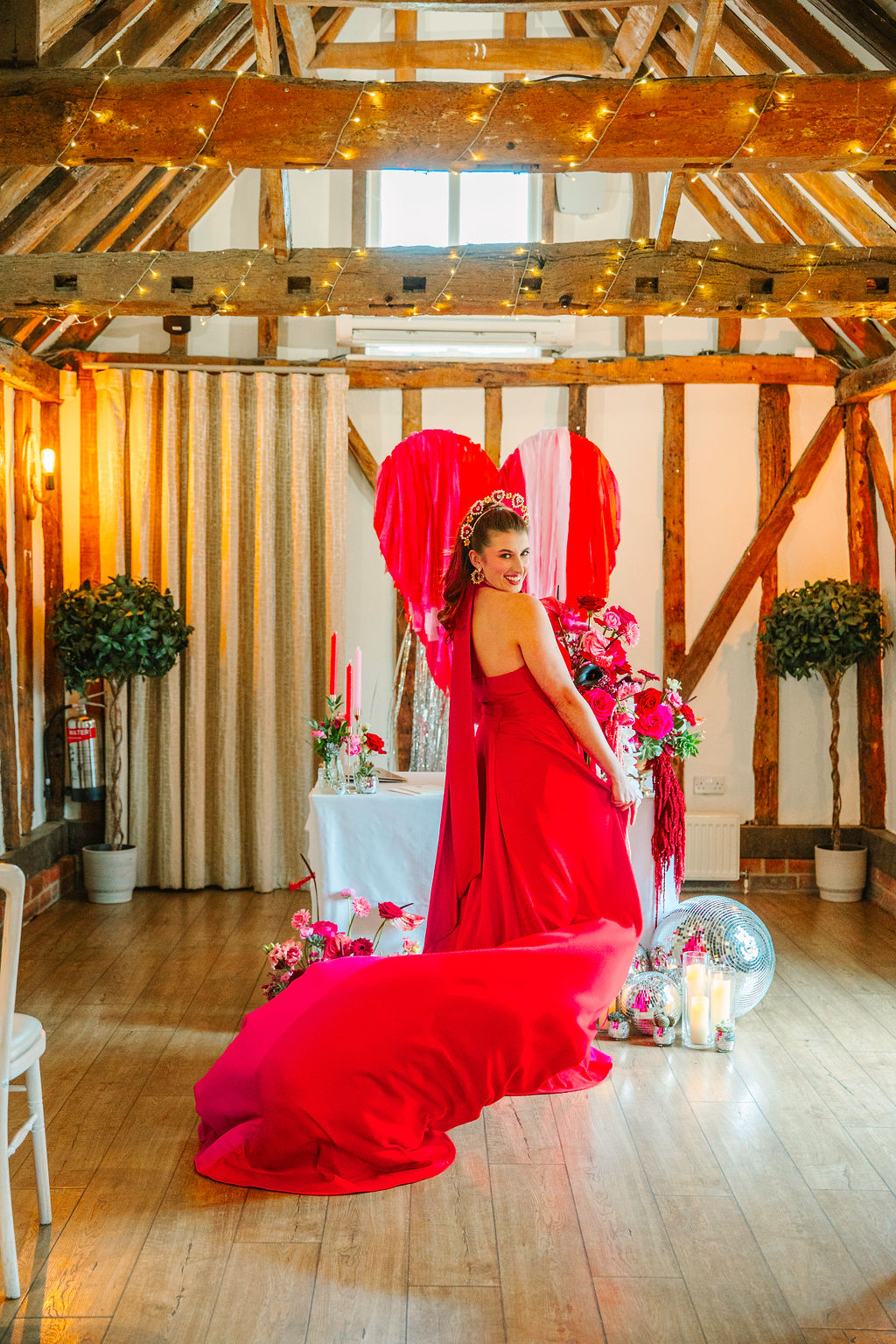 Bride wearing a pink and red wedding dress turning in front of the wedding ceremony area within the fairy lit beamed barn wedding venue.
