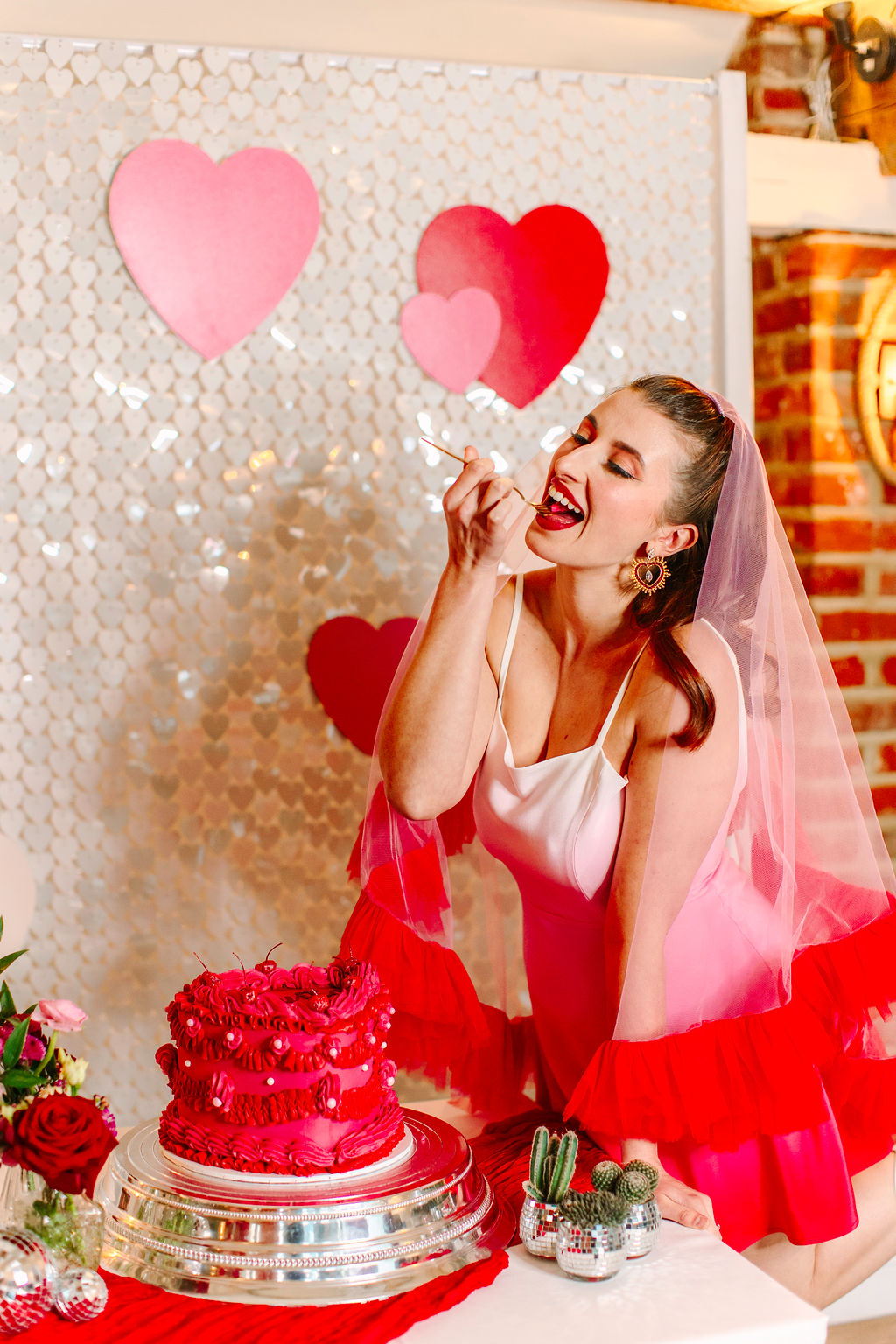 Bride wearing pink and red veil and red and pink dip dye mini wedding dress leans on the wedding cake table feeding herself a slice