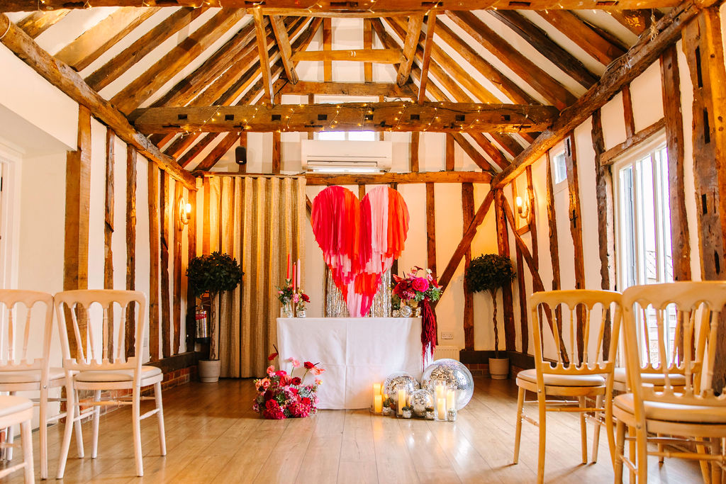 Rustic beamed wedding venue with pink and red love heart ceremony backdrop, disco balls and statement wedding flowers.