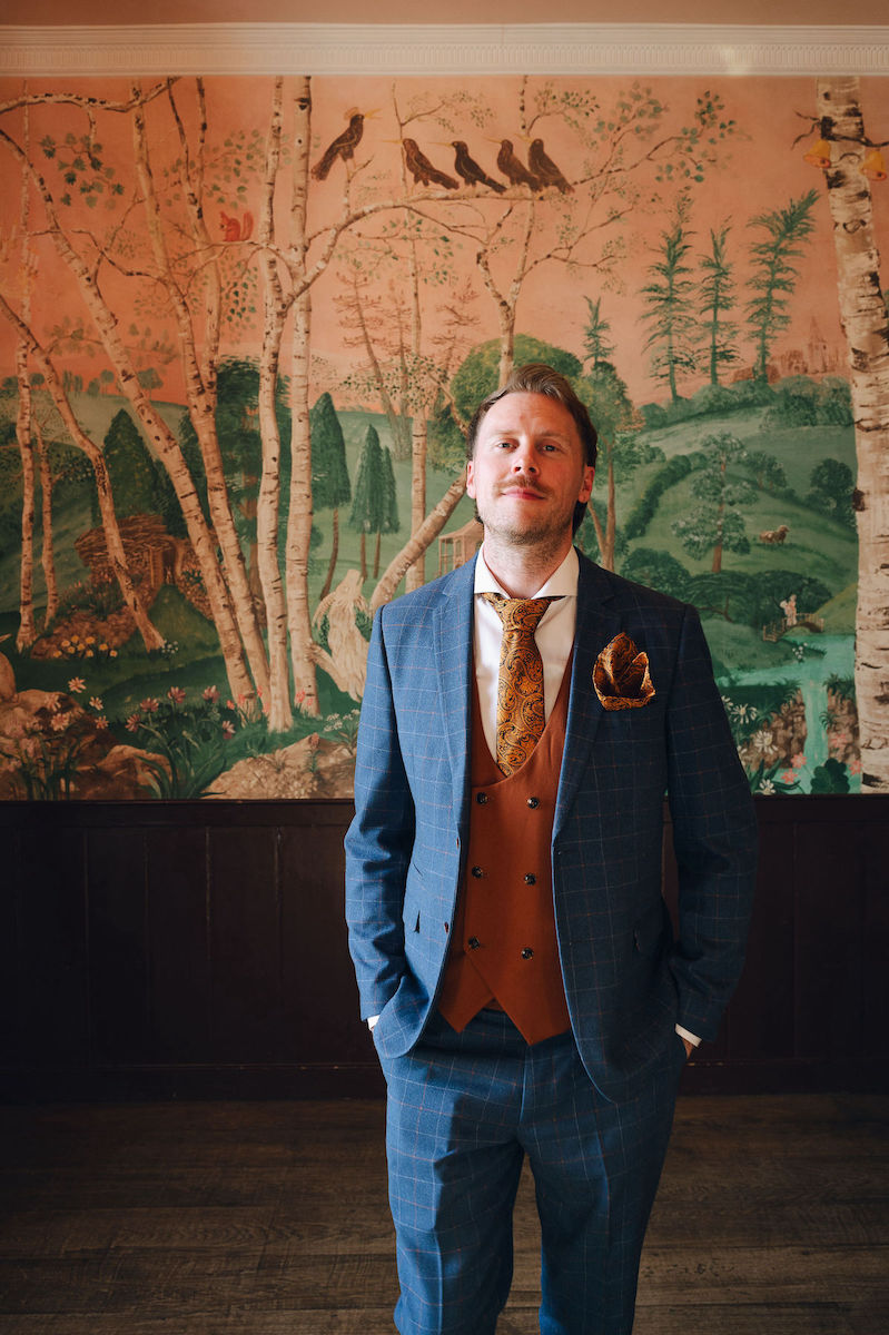 Groom stood in the alternative wedding venue with statement feature all wearing blue suit with orange waistcoat and orange mustard paisley tie.