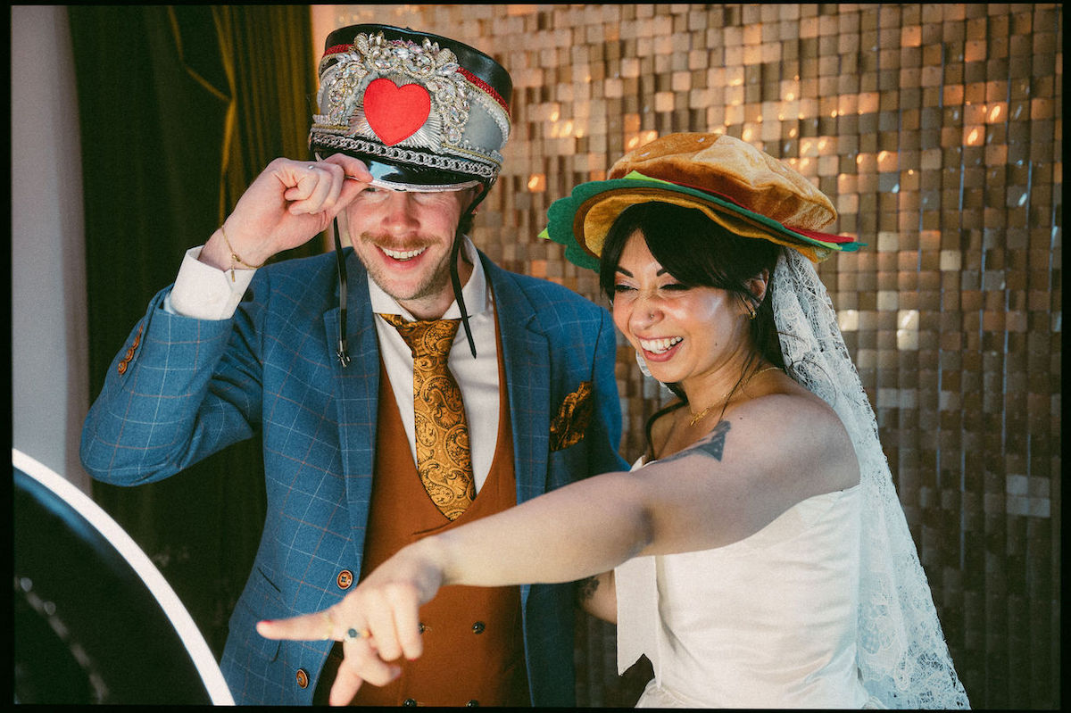 Bride pointing at a screen while she and the groom have fun with silly hats at wedding photo booth.