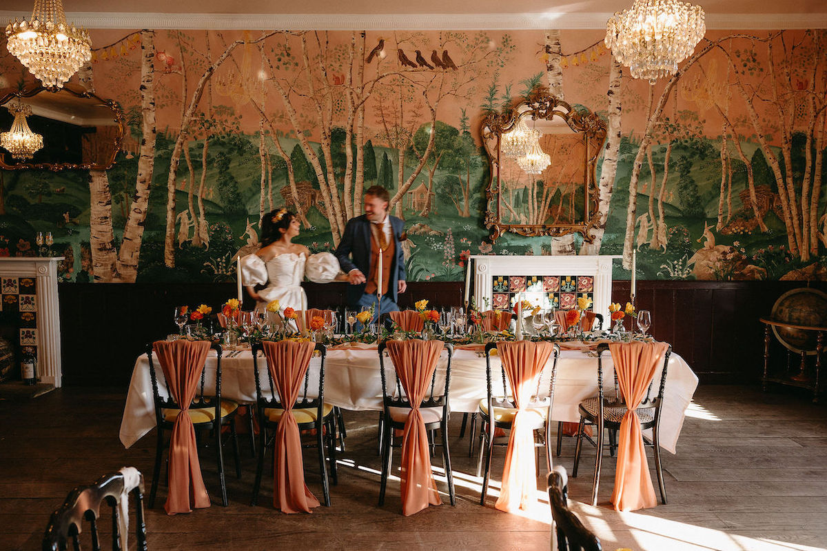 couple entering the wedding breakfast room for cool retro wedding. Table styled with orange and yellow flowers and chair covers. Bride and groom wear retro wedding outfits