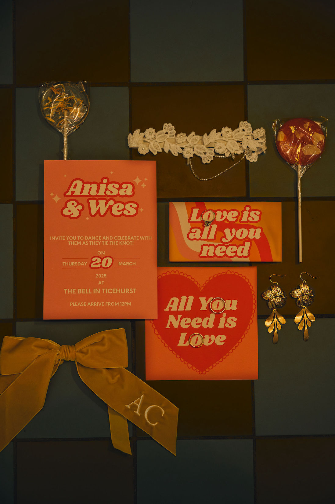 Flat lay with alternative wedding stationery in orange and peach with retro waves, hearts and stars. Monogram bow place setting, vintage earrings, floral garter and fun lollypops are set around the stationery.