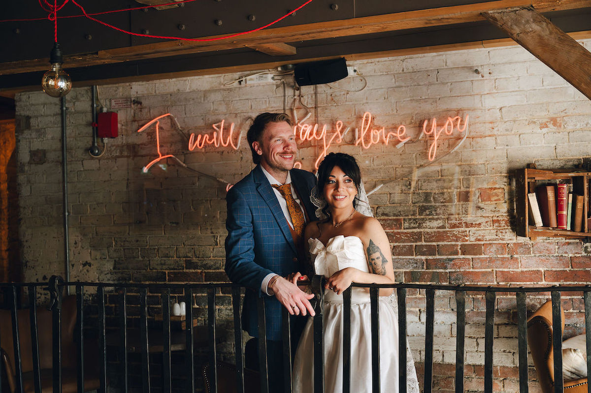 Bride and groom stood in front of a neon sign saying 'I will always love you' on retro music inspired wedding day