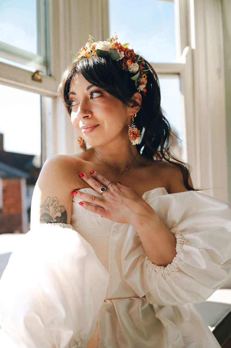 Bride in the window of the room she has got ready in wearing modern puff sleeve wedding dress, dried flower crown and dried flower earrings