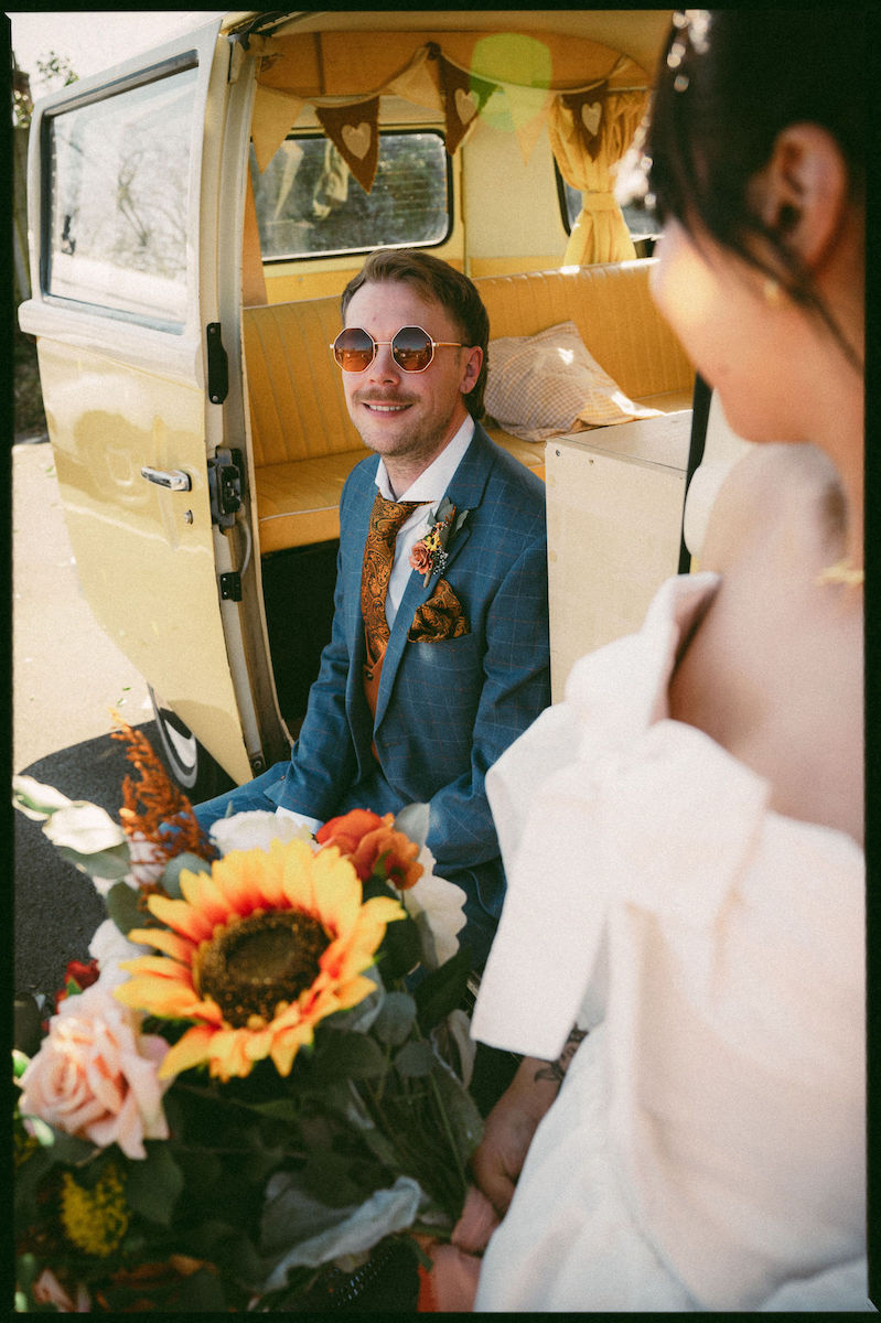 Groom sitting in the door of a vintage VW camper van wearing 70s sunglasses, blue suit and paisley tie
