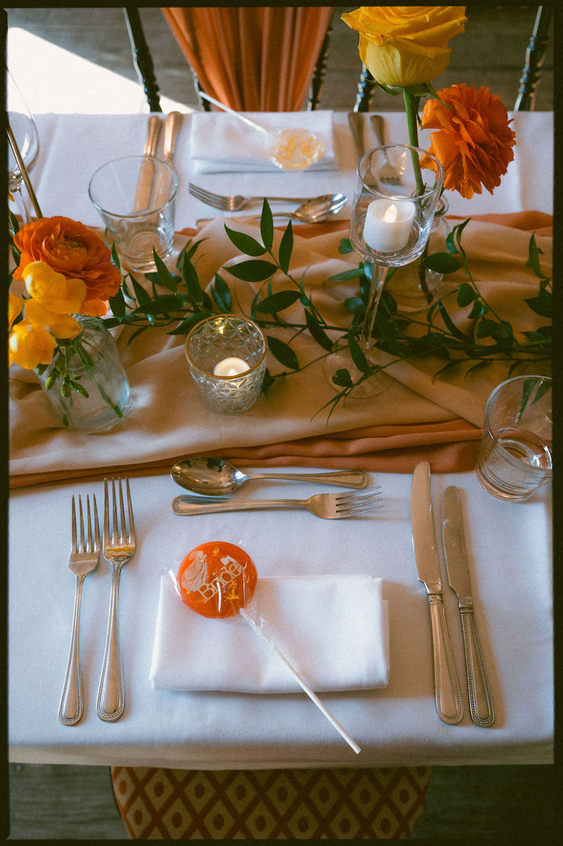 Bride place setting lolly on top of a napkin on the wedding breakfast table dressed with orange runners, candles and orange and yellow flowers in bud vases