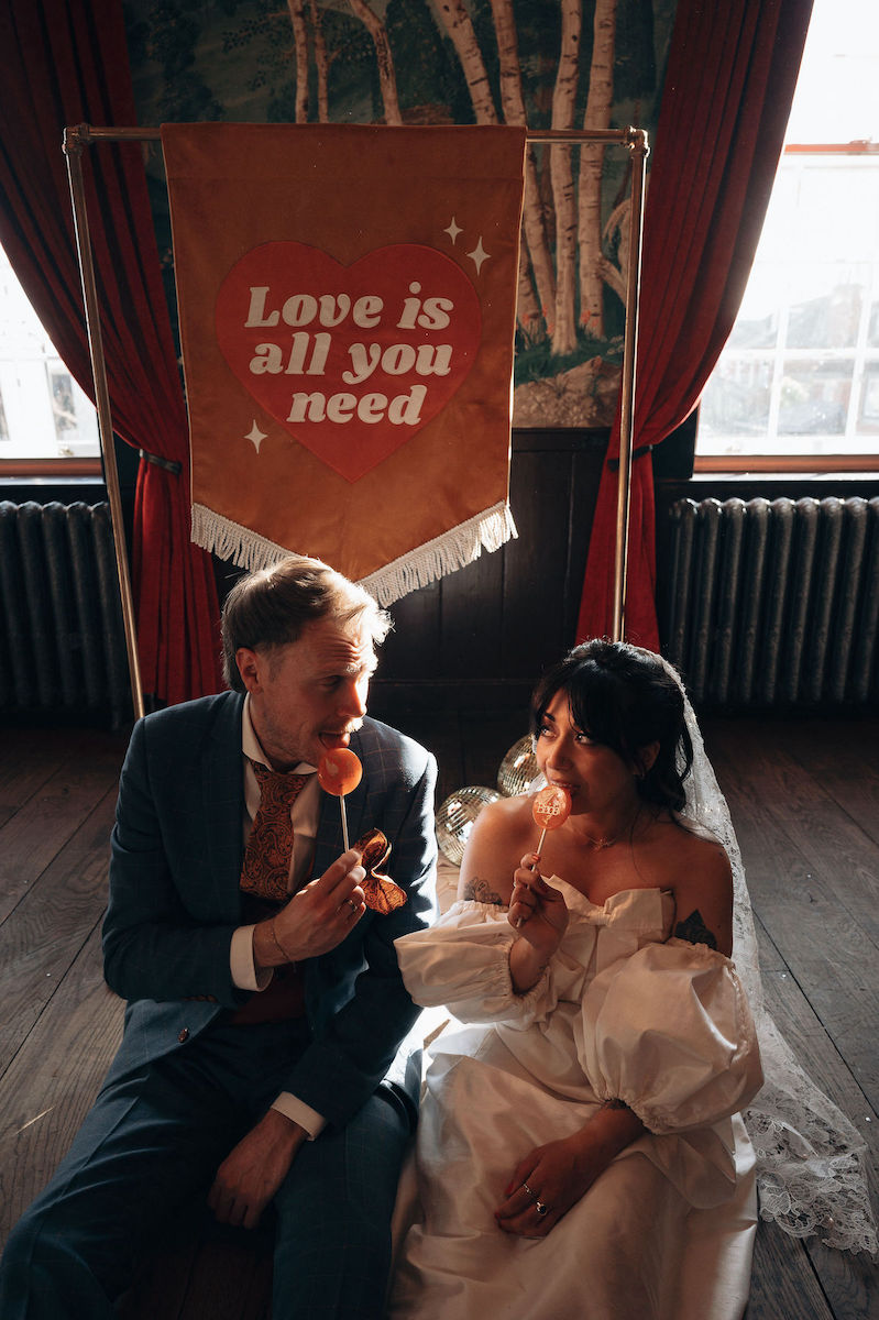 Bride and groom sit in the floor in front of a 'love is all you need' wedding banner. The couple are licking orange personalised place setting lollies.