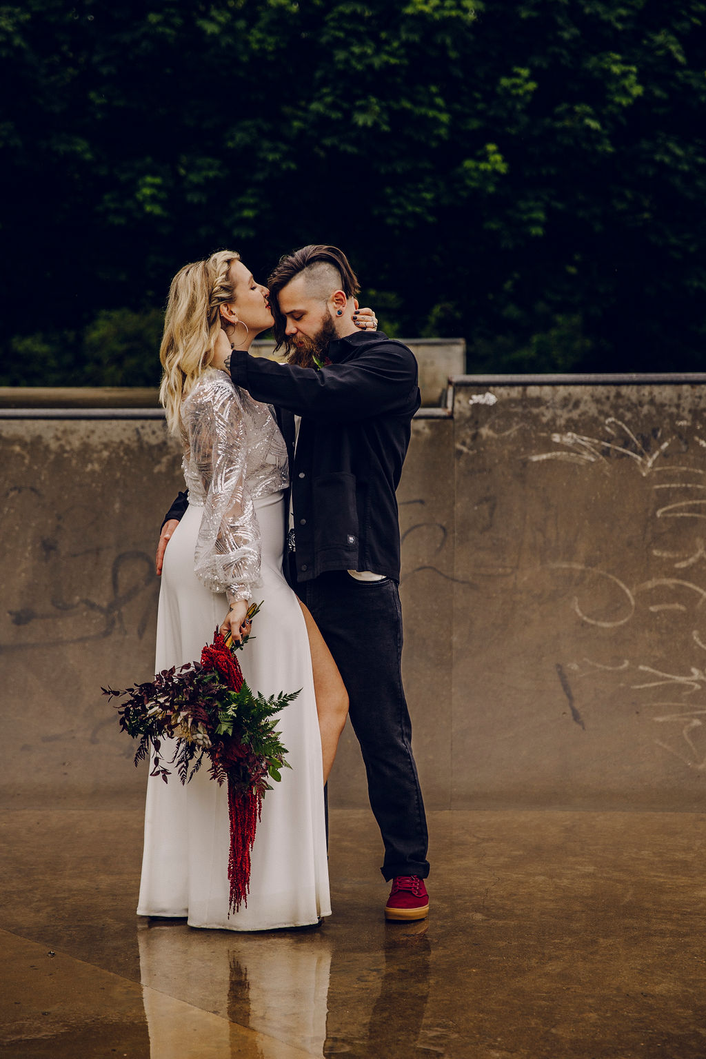 Alternative emo couple embracing in front of urban skate park ramp. The bride wears a wedding dress and the groom jeans and jacket
