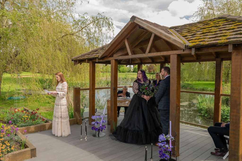 Bride and groom in outdoor wedding ceremony stood by a lake. A lady in cream is doing a reading