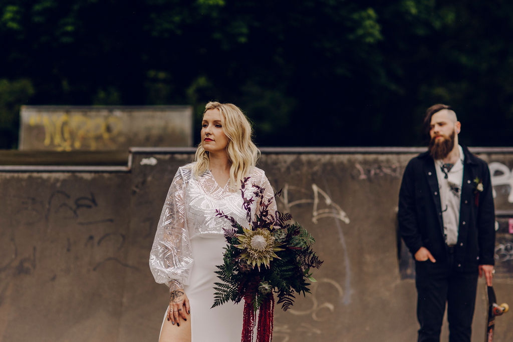 bride stood in focus holding an alternative wedding bouquet. Groom with mohawk in the background