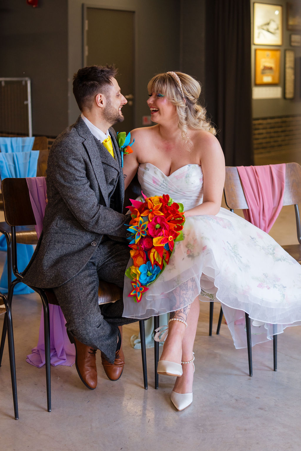 bride and groom sit on wedding ceremony chairs covered with rainbow sashes. The bride holds a vibrant fabric flower bouquet.