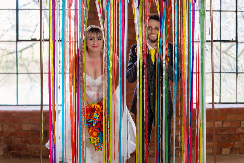 bride and groom peek out from handmade bright ribbon ceremony backdrop
