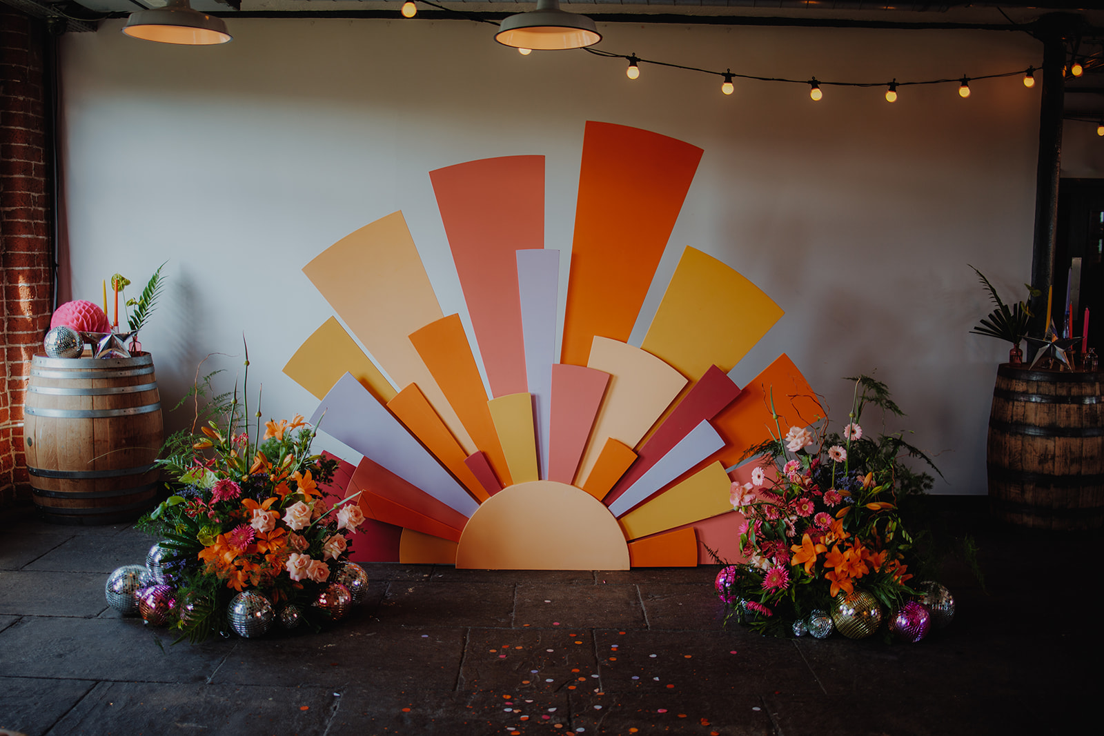 statement ceremony backdrop - a radiating sun in pink, red, orange, yellow and lilac. Statement floral arrangements sit either side.