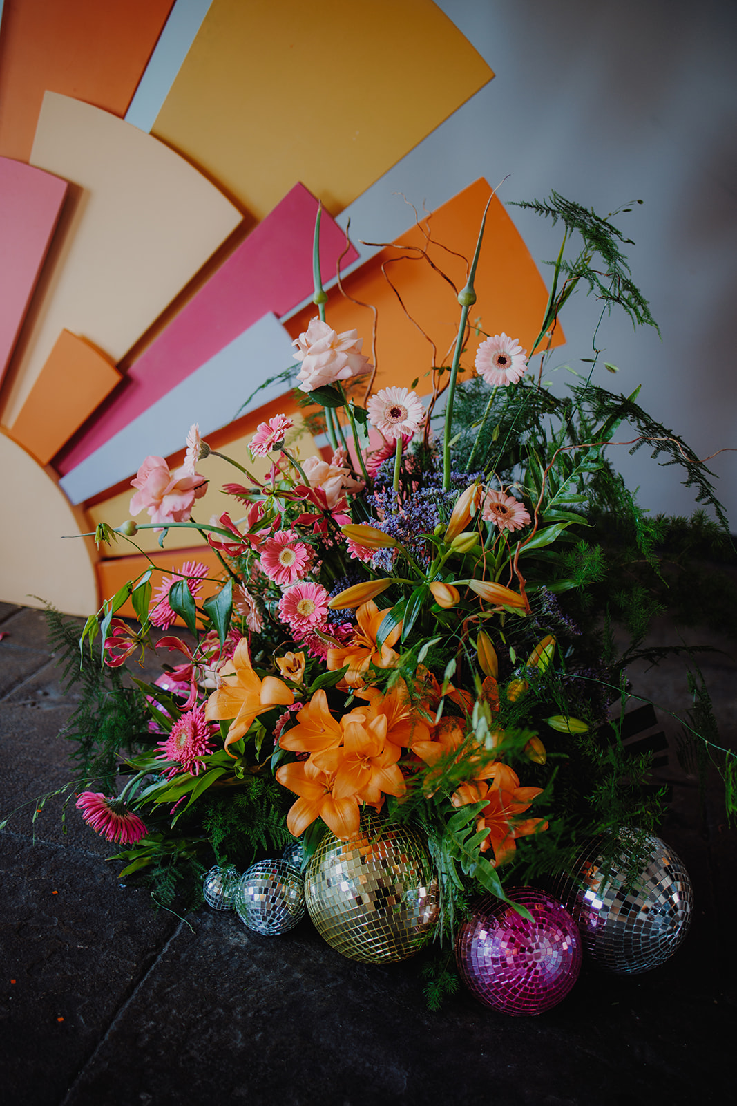 striking floor display of flowers in orange, pink and purple with disco balls around the bottom. Set to the side of a sun beam ceremony backdrop