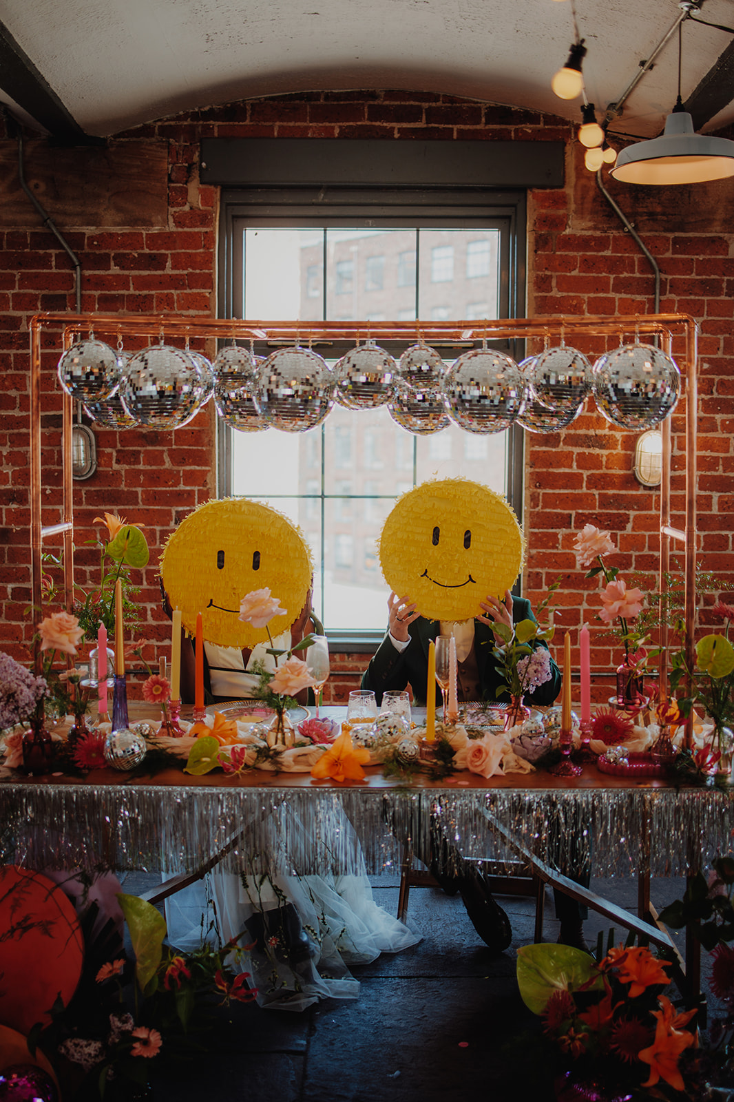 bride and groom hold up smiley face piñatas sat at disco ball themed wedding breakfast table