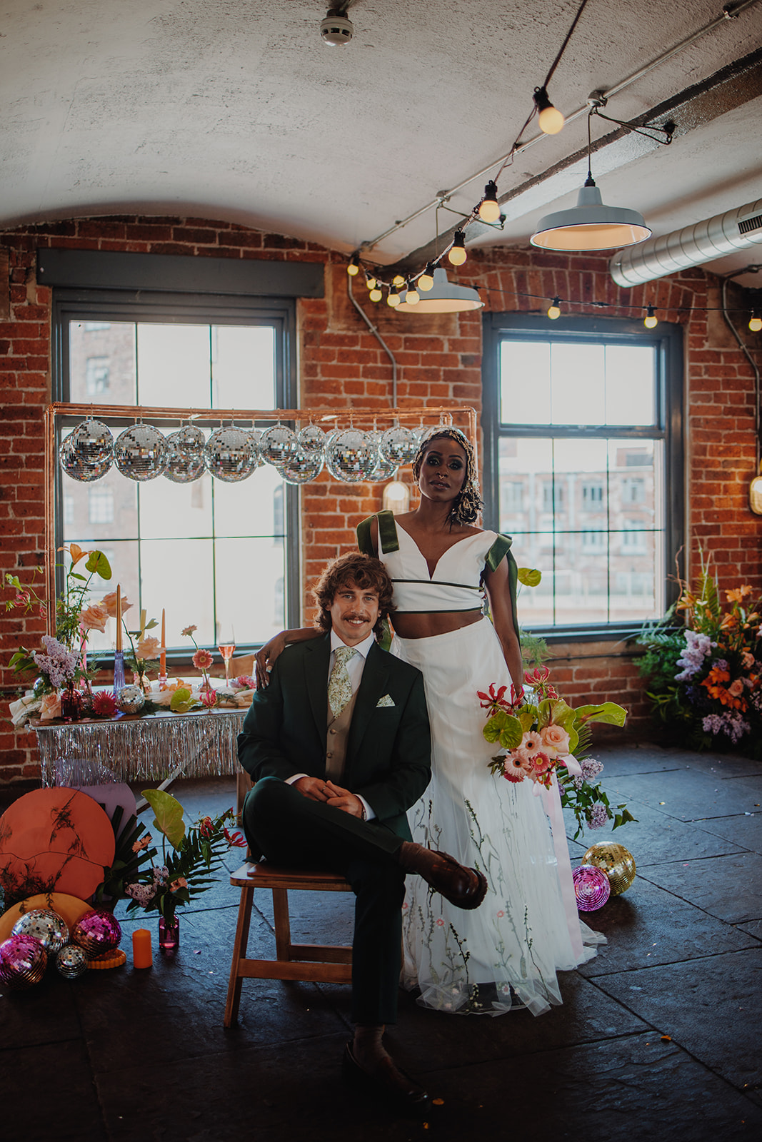 POC bride wearing wedding separates stands with groom in green suit. A playful vibrant wedding breakfast table with disco ball canopy is behind them